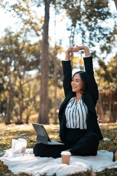 Happy millennial businesswoman sitting on grass with laptop in the park and stretching arms.