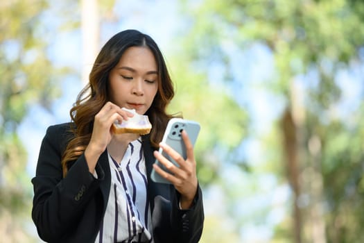 Young businesswoman using mobile phone and eating tasty sandwiches during lunch.