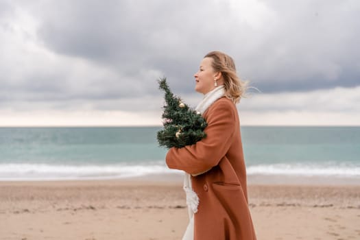 Blond woman Christmas sea. Christmas portrait of a happy woman walking along the beach and holding a Christmas tree on her shoulder. She is wearing a brown coat and a white suit