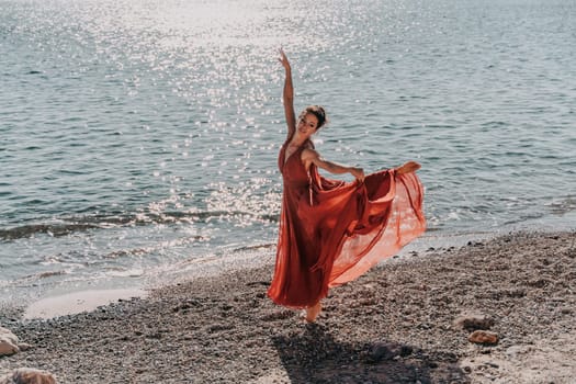 Woman red dress sea. Female dancer in a long red dress posing on a beach with rocks on sunny day. Girl on the nature on blue sky background