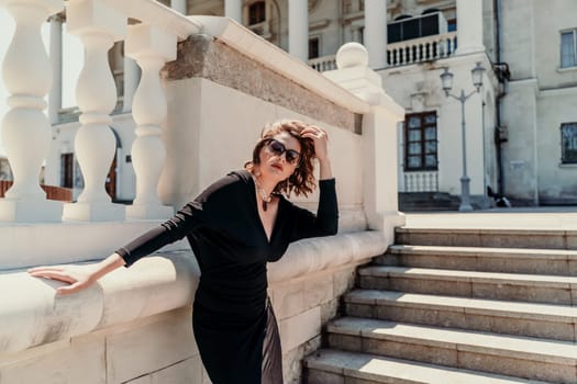 Stylish woman in the city. Fashion photo of a beautiful model in an elegant black dress posing against the backdrop of a building on a city street.