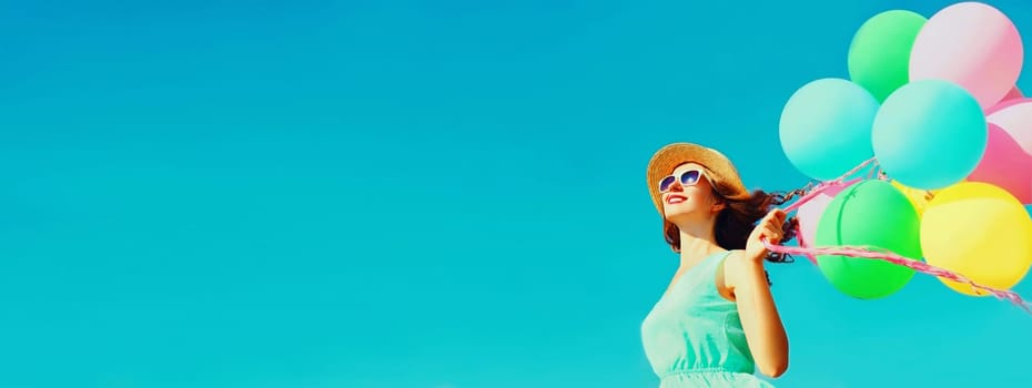 Happy smiling young woman with bunch of colorful balloons wearing a summer straw hat on the field on a blue sky background