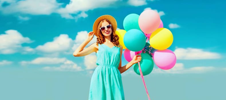 Happy smiling young woman with bunch of colorful balloons wearing a summer straw hat on the field on a blue sky background