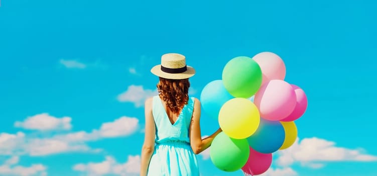 Rear view of young woman with bunch of colorful balloons wearing summer straw hat on the field on blue sky background