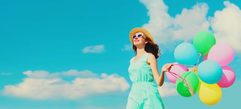 Happy smiling young woman with bunch of colorful balloons wearing a summer straw hat on the field on a blue sky background