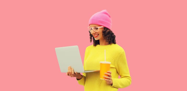 Stylish modern happy young woman working with laptop wearing colorful clothes, yellow sweater, hat and eyeglasses on pink studio background
