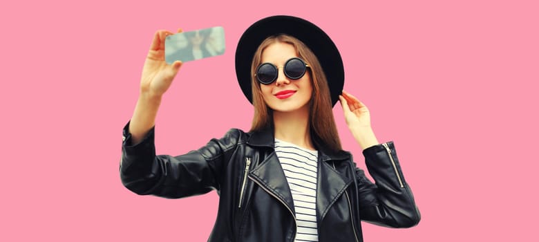 Portrait of beautiful young woman taking selfie with smartphone posing wearing black round hat on pink background