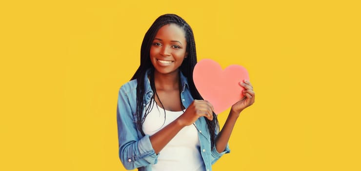Portrait of happy smiling young african woman holding paper red heart on colorful yellow background