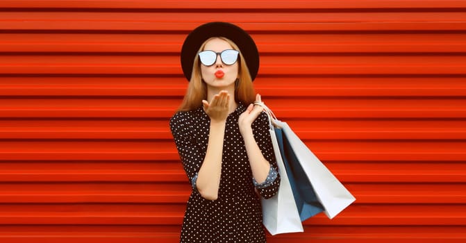 Portrait of beautiful young woman with shopping bags blowing kiss on red background