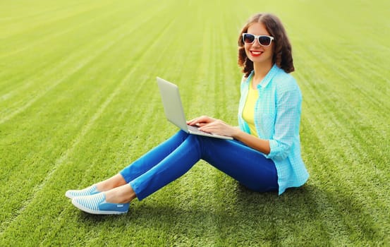 Happy young woman working with laptop in summer park sitting on green grass background