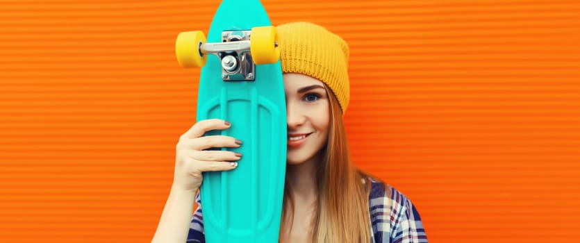 Portrait of modern smiling young woman with green skateboard wearing colorful yellow hat on orange background