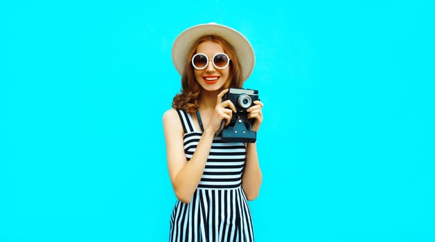 Summer portrait of happy smiling young woman photographer with vintage film camera wearing straw hat, dress, sunglasses on blue background