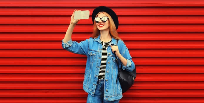 Portrait of happy smiling young woman taking selfie with smartphone wearing jean jacket, black round hat on red background