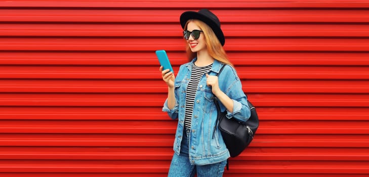 Portrait of stylish modern smiling young woman with smartphone wearing jean jacket, black round hat on red background