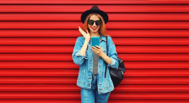 Portrait of stylish modern smiling young woman with smartphone wearing jean jacket, black round hat on red background