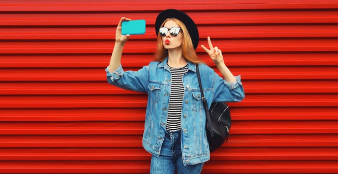 Portrait of beautiful young woman blowing her lips sends kiss taking selfie with smartphone wearing jean jacket, black round hat on red background