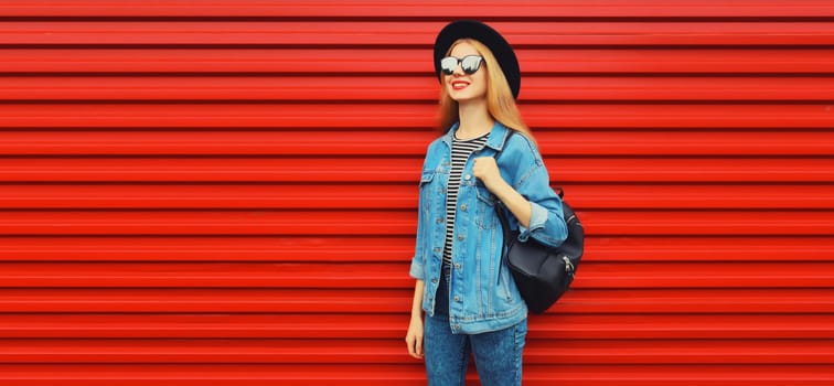 Portrait of happy smiling young woman looking away wearing black round hat, jean jacket, backpack on red background, blank copy space for advertising text