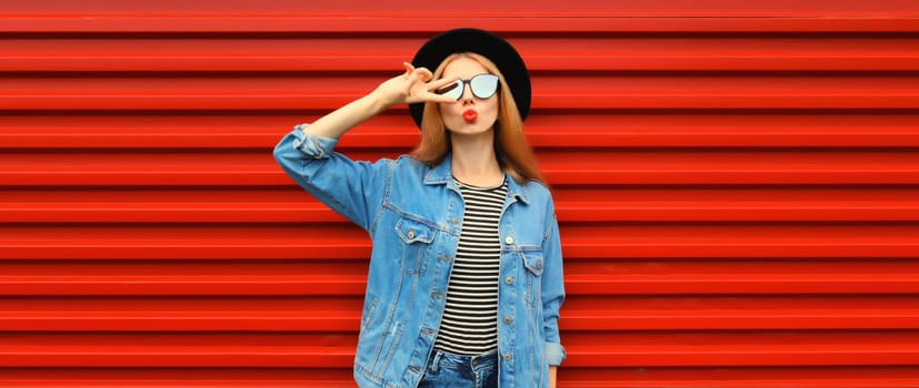 Portrait of stylish young woman model blowing her lips sending sweet air kiss wearing jean jacket, black round hat on red background