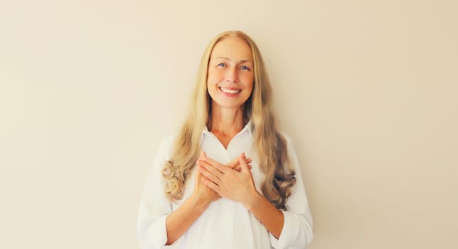 Portrait of beautiful happy smiling caucasian middle aged woman put folded hands on her heart looking at camera on white studio background