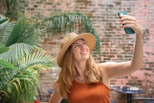Woman wearing a sun hat is smiling and gesturing while taking a selfie with her cell phone in a leisurely travel landscape surrounded by terrestrial plants. She looks happy in the sunny weather