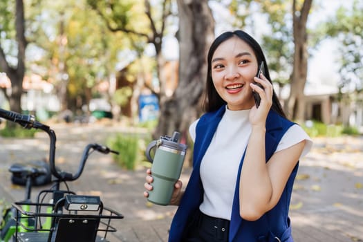 Smiling asian businesswoman hold reusable eco-friendly ecological cup and using mobile while sitting at park.