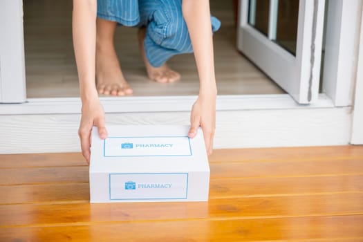 Close up hands of sick Asian woman sitting at door to receive medication first aid pharmacy box from hospital delivery service at floor home, female patient buy drugstore online business, healthcare