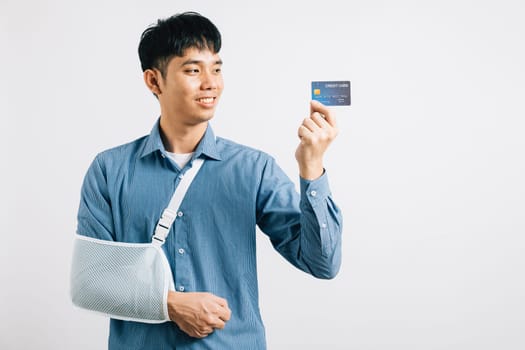 Injured man confidently smiles with a damaged arm in a support splint, managing medical expenses via credit card. Happy Asian man with sling support hand, isolated on white, showcasing health care.