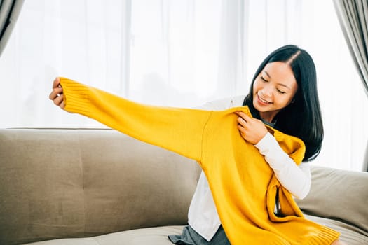 Happy Asian woman reveals a shirt from a box smiling with delight at home. Satisfied shopper unboxes online order unveiling new clothes. Delivery and home shopping concept.