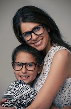Happy mother, portrait and hugging daughter with love for care or support in fashion on a gray studio background. Mom, child or young kid with smile and glasses in happiness for parenting or bonding.