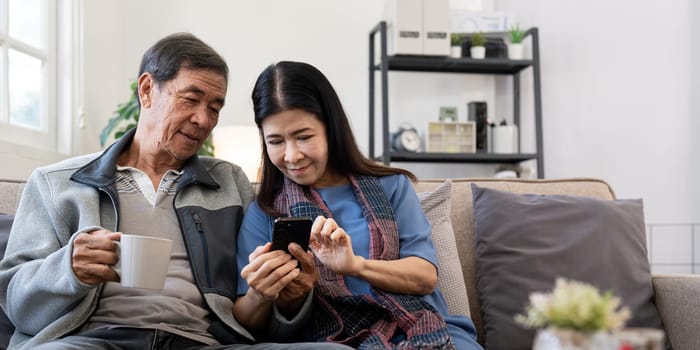 Happy elderly asian couple using smartphone sit on sofa doing ecommerce shopping online on website at home.