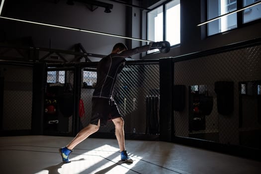 Male athlete in sportswear and boxing gloves shadowboxing, highlighted by rays of sunlight in well equipped MMA cage in dimly lit training room