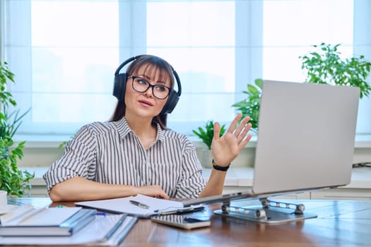 Middle-aged woman in headphones working at computer in home office, looking at web cam talking. Online meeting, remote work teaching financial legal advice interview blogging freelance, mental therapy