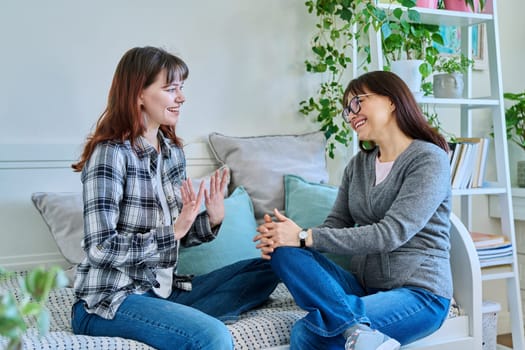 Talking happy smiling mother and teenage daughter sitting together on couch at home. Family, communication, motherhood, friendship, relationship between parent and daughter 18-20 years old