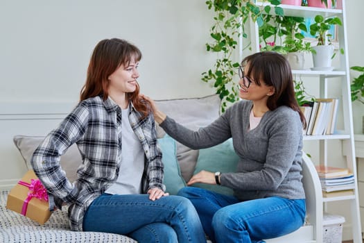 Cheerful teenage daughter congratulating middle aged mother with gift, sitting together on couch at home. Holiday birthday, congratulations, love, mother's day, happiness joy concept