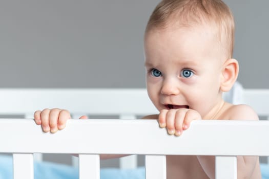 A happy toddler in a white crib is standing and looking over the railing with a smile on its face