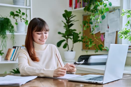 Young female college student studying at home at desk using computer laptop, writing in notebook. E-learning, education, technology, knowledge, youth concept