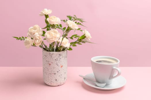 Bouquet of white carnations in a vase and a cup of coffee on the pink background.