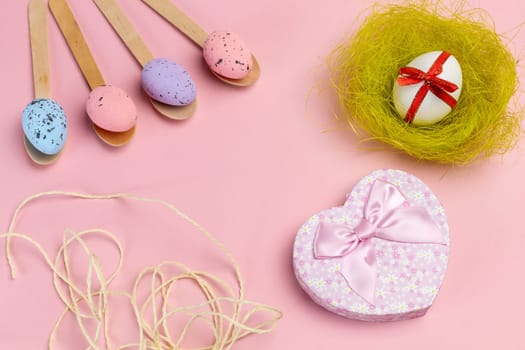Colored Easter eggs on wooden spoons, a nest, a rope and a gift box on the pink background. Top view.