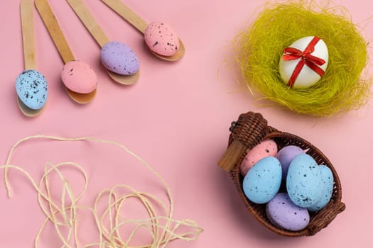 Colored Easter eggs on wooden spoons and in a wicker basket with a nest on the pink background. Top view.