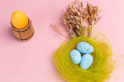 Nest with colored Easter eggs on the pink background with decor plants and wooden utensil. Top view.