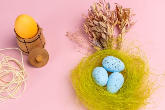 Nest with colored Easter eggs on the pink background with decor plants and wooden utensil. Top view.