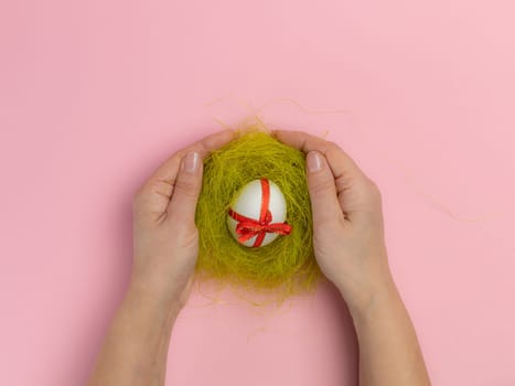 Woman is holding a nest with the Easter egg tied with a red ribbon. Top view. Pink background.