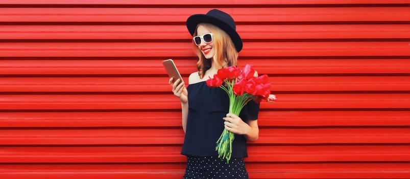 Beautiful smiling woman with smartphone holding bouquet of red rose flowers in black round hat