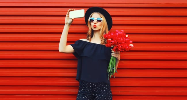 Beautiful young woman taking selfie with smartphone holding bouquet of red rose flowers in black round hat