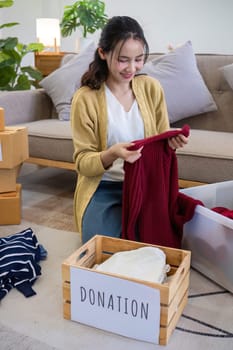 Cute Asian woman sitting next to sofa in living room sorting unwanted clothes for donation..