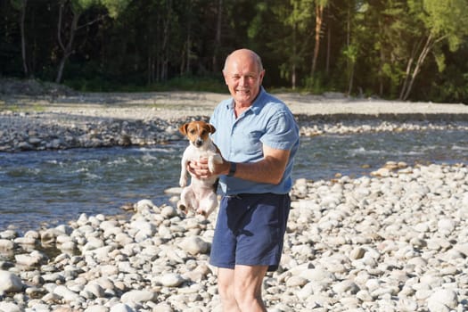Senior man holding small wet Jack Russell terrier dog in his hand, after she swam in the near river on sunny day