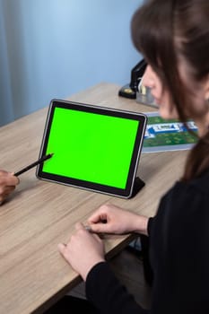 A businesswoman is sitting at her desk and using a tablet with a green screen. She is smiling and looking at the tablet.