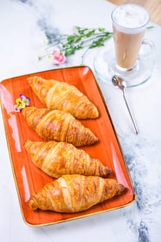 A delightful breakfast with croissants and coffee on a marble table. Croissants are golden brown on an orange plate, coffee with froth.