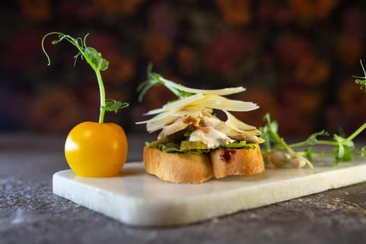 A bruschetta with chicken, pesto, and parmesan on a white marble board. The bruschetta is garnished with microgreens and a pea pod.