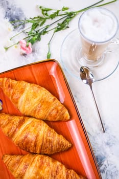 A delightful breakfast with croissants and coffee on a marble table top view. Croissants are golden brown on an orange plate, coffee with froth.